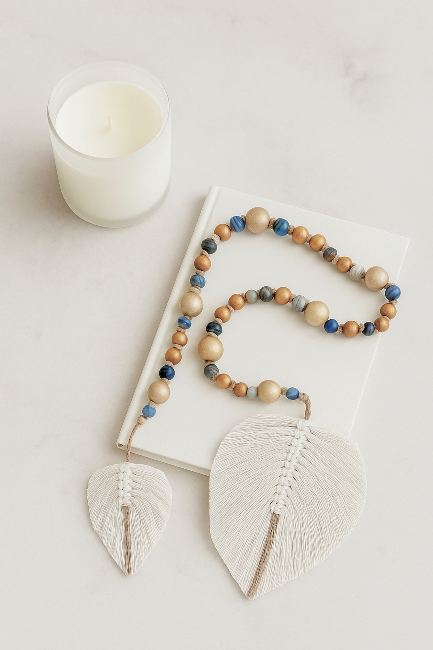 Handmade boho garland made of natural wood and glass beads in earthy blue tones, finished with two white fiber leaf charms, styled on a white book next to a minimal candle.