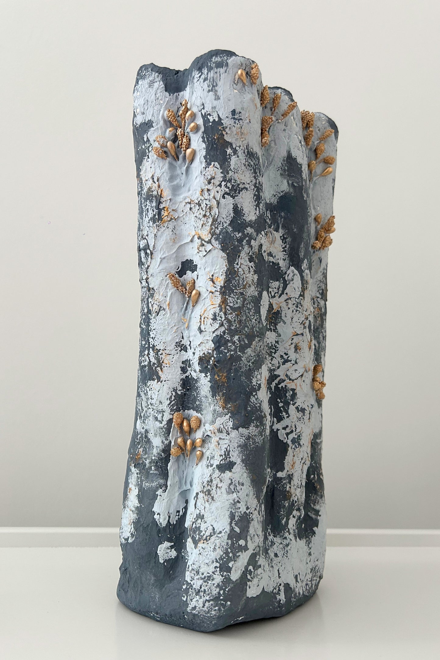 Organic tall vase with grey-blue plaster texture and gold accent details, standing against white wall.