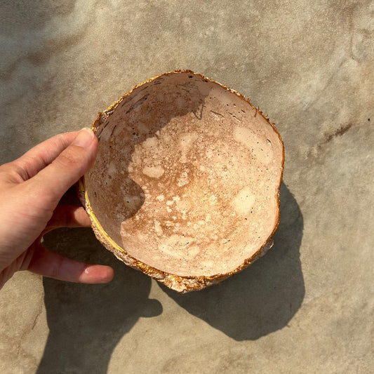 Handmade rustic bowl with gold detailing, photographed on a warm marble surface in sunlight.