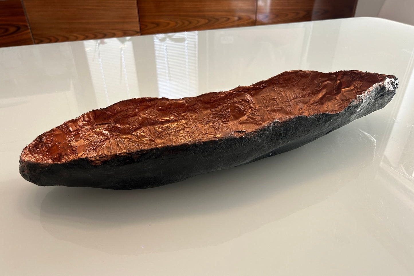 Sculptural long bowl with copper interior and matte black exterior placed on rustic wood.