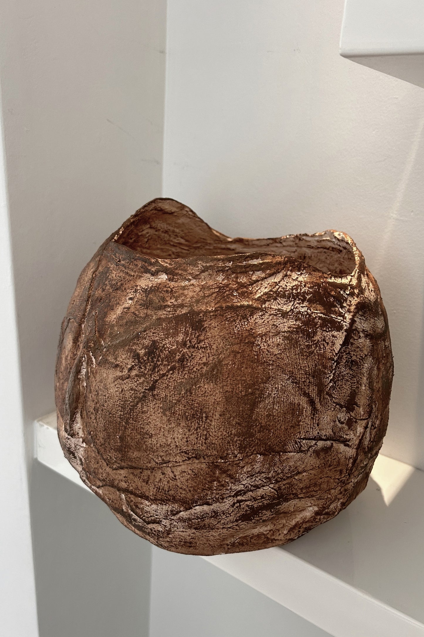 Side angle of a rustic handmade vase showing natural texture and aged bronze tones on a white shelf.