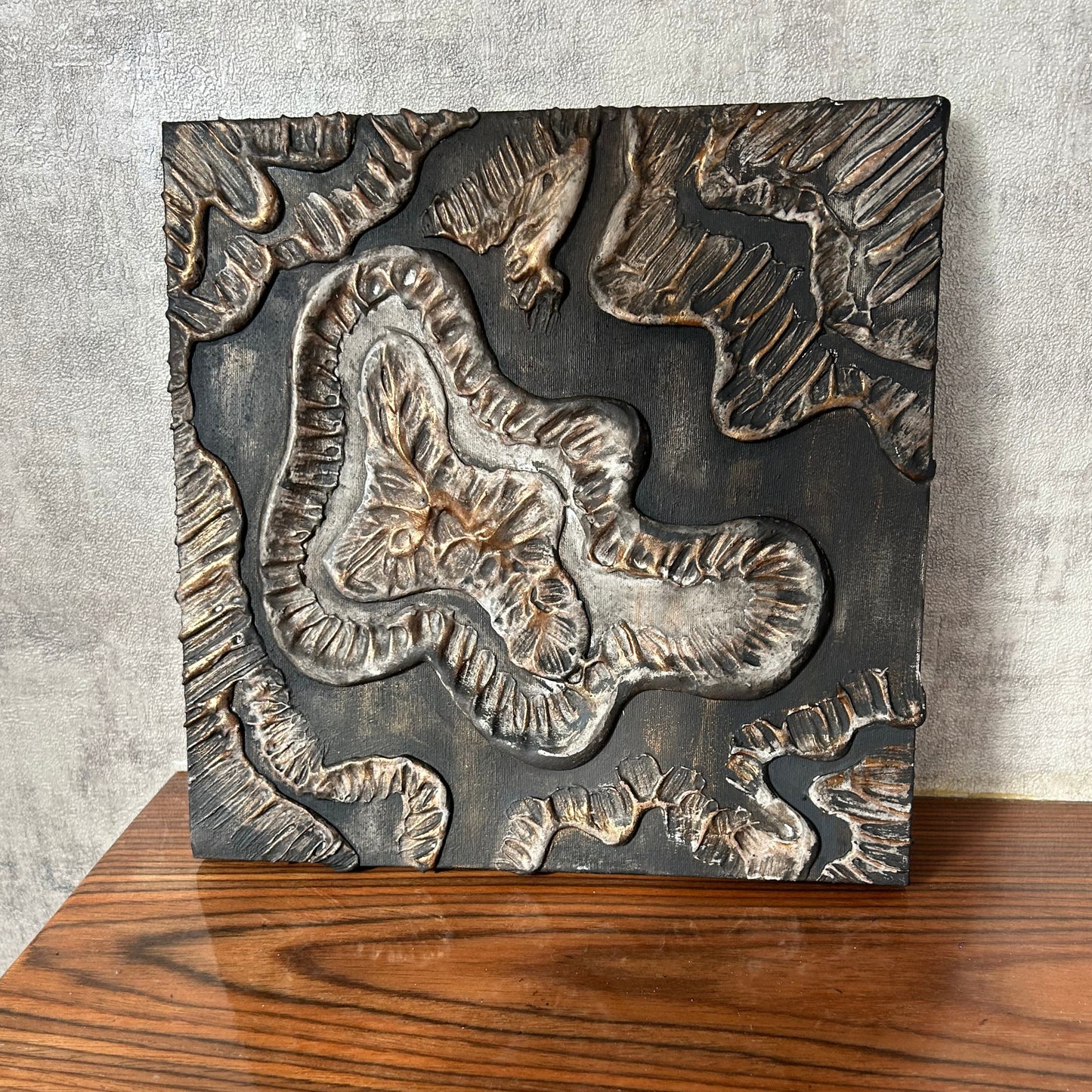 Handmade sculptural wall panel resting on wooden base, bronze and charcoal colors
