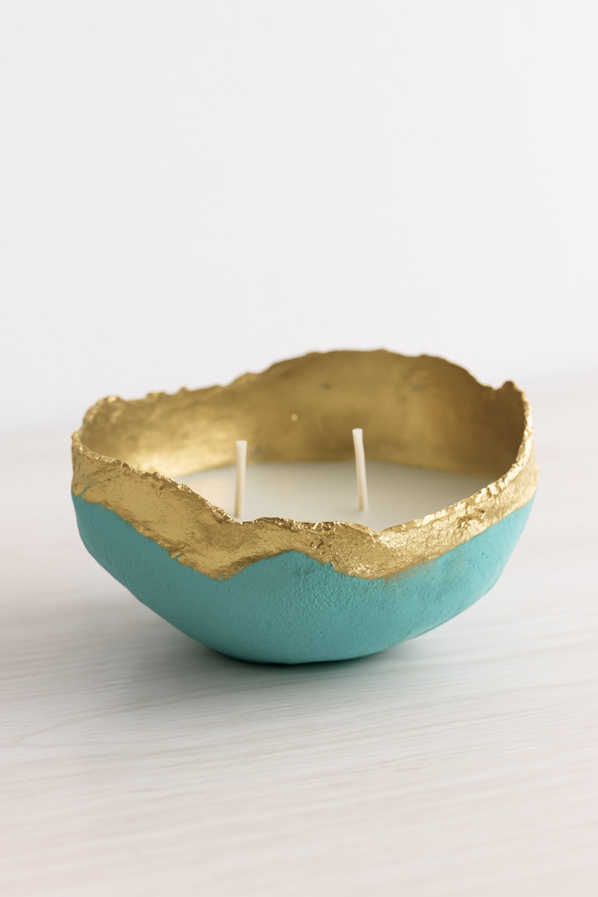 Close-up of a turquoise handmade candle bowl with shimmering gold inner rim and double wicks, placed on a light wood surface.