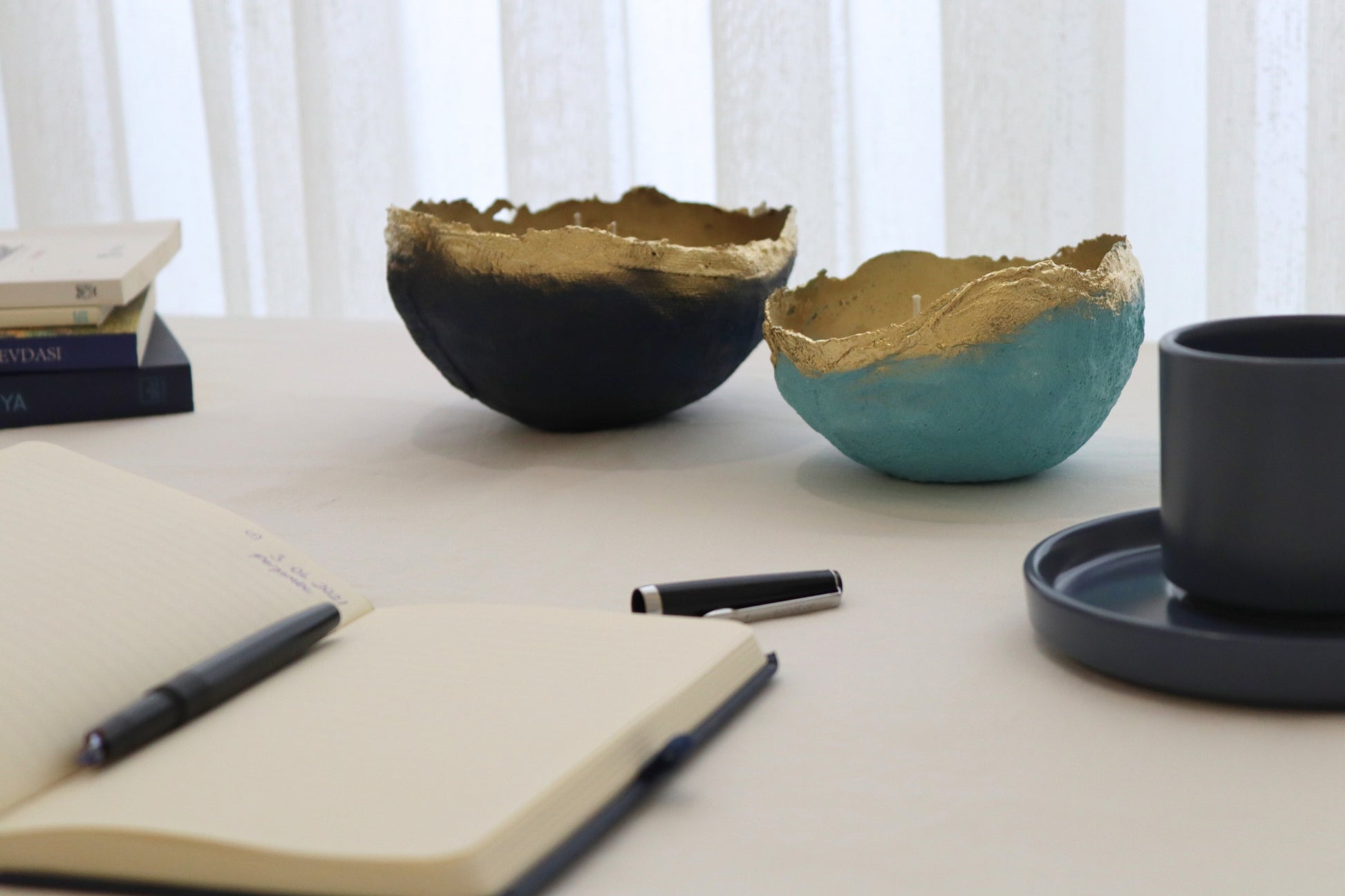 Minimalist table styling featuring handmade candle bowls with gold edges, next to books and a cup.
