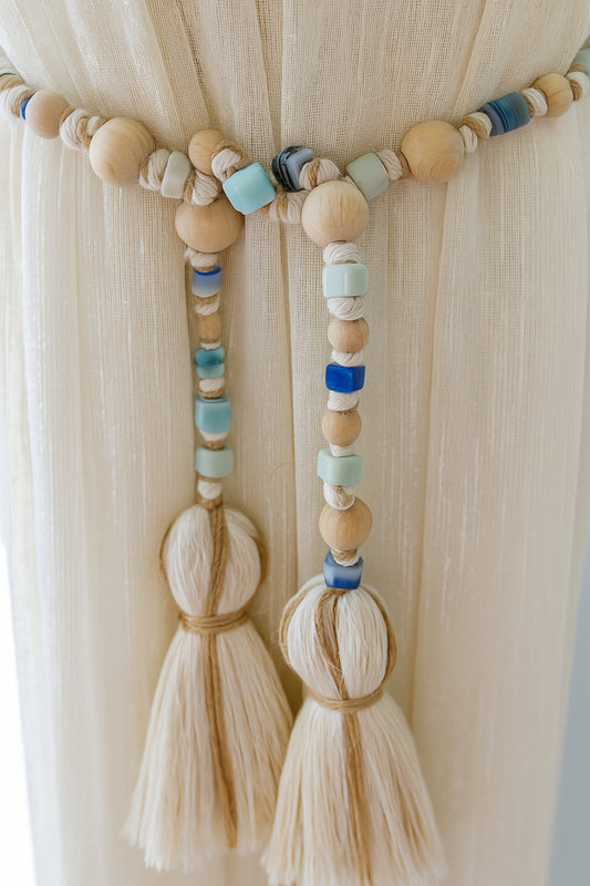 Boho-style curtain tieback garland with natural wooden and blue glass beads hanging on sheer white curtains.
