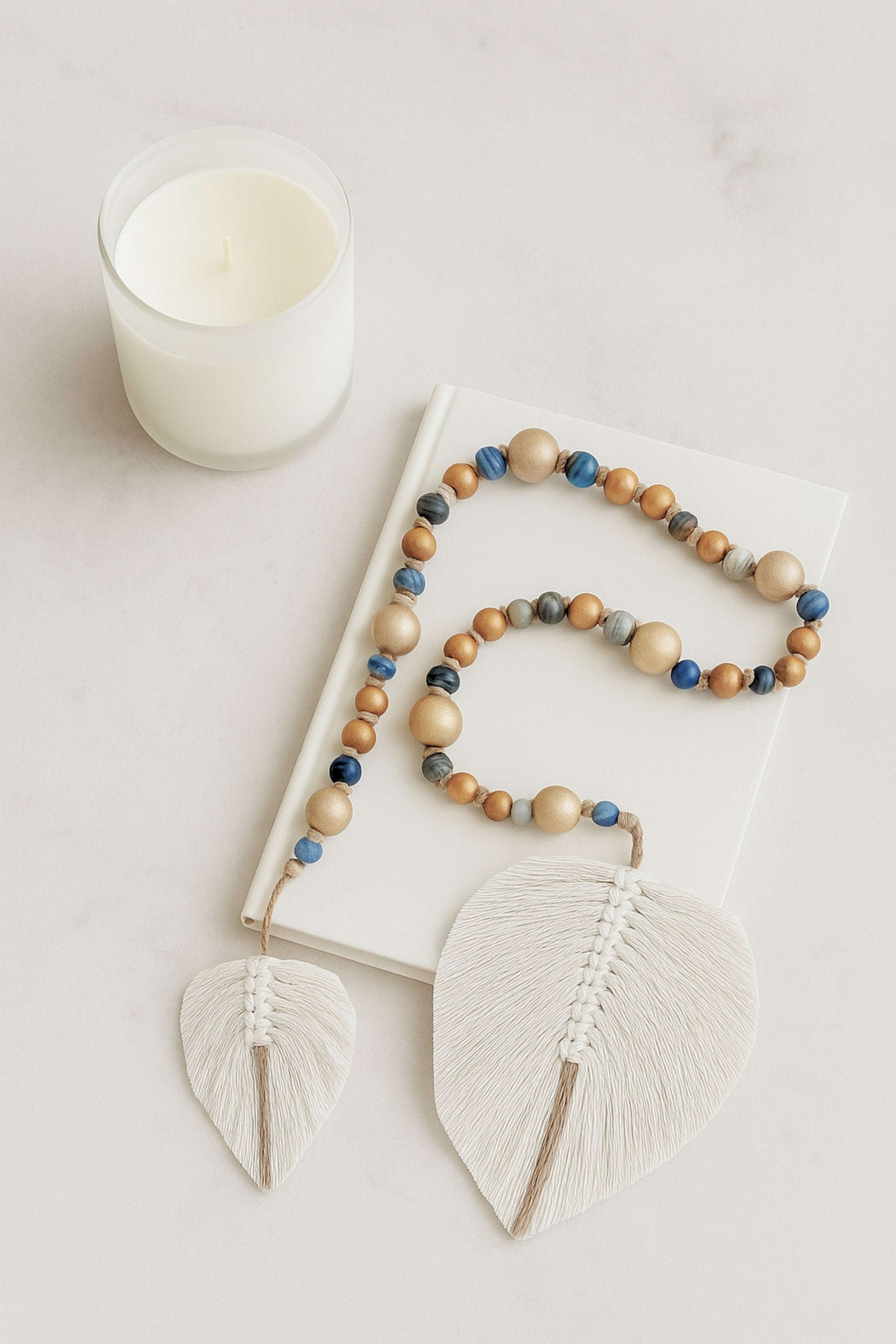 Handmade boho garland made of natural wood and glass beads in earthy blue tones, finished with two white fiber leaf charms, styled on a white book next to a minimal candle.