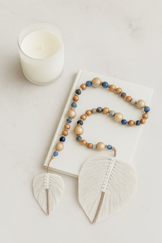 Handmade boho garland made of natural wood and glass beads in earthy blue tones, finished with two white fiber leaf charms, styled on a white book next to a minimal candle.