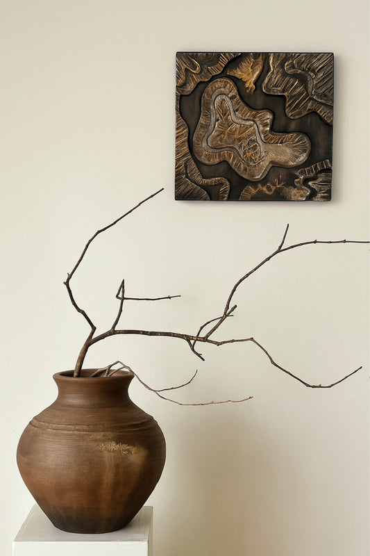Handmade bronze and black sculptural wall art displayed above a rustic earthen vase with natural dried branch, styled in a minimalist Japandi interior
