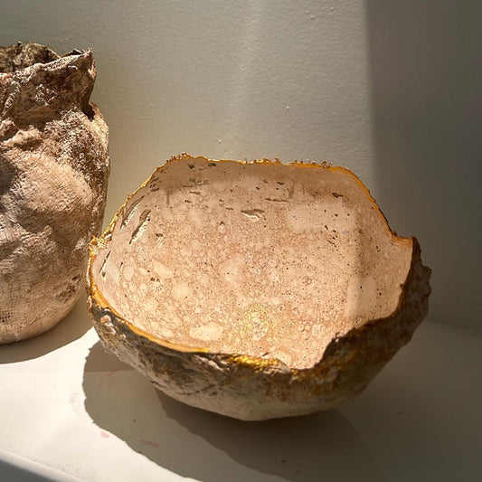 Gold-edged bowl placed on a white shelf beside another handmade vessel, sunlit corner view.