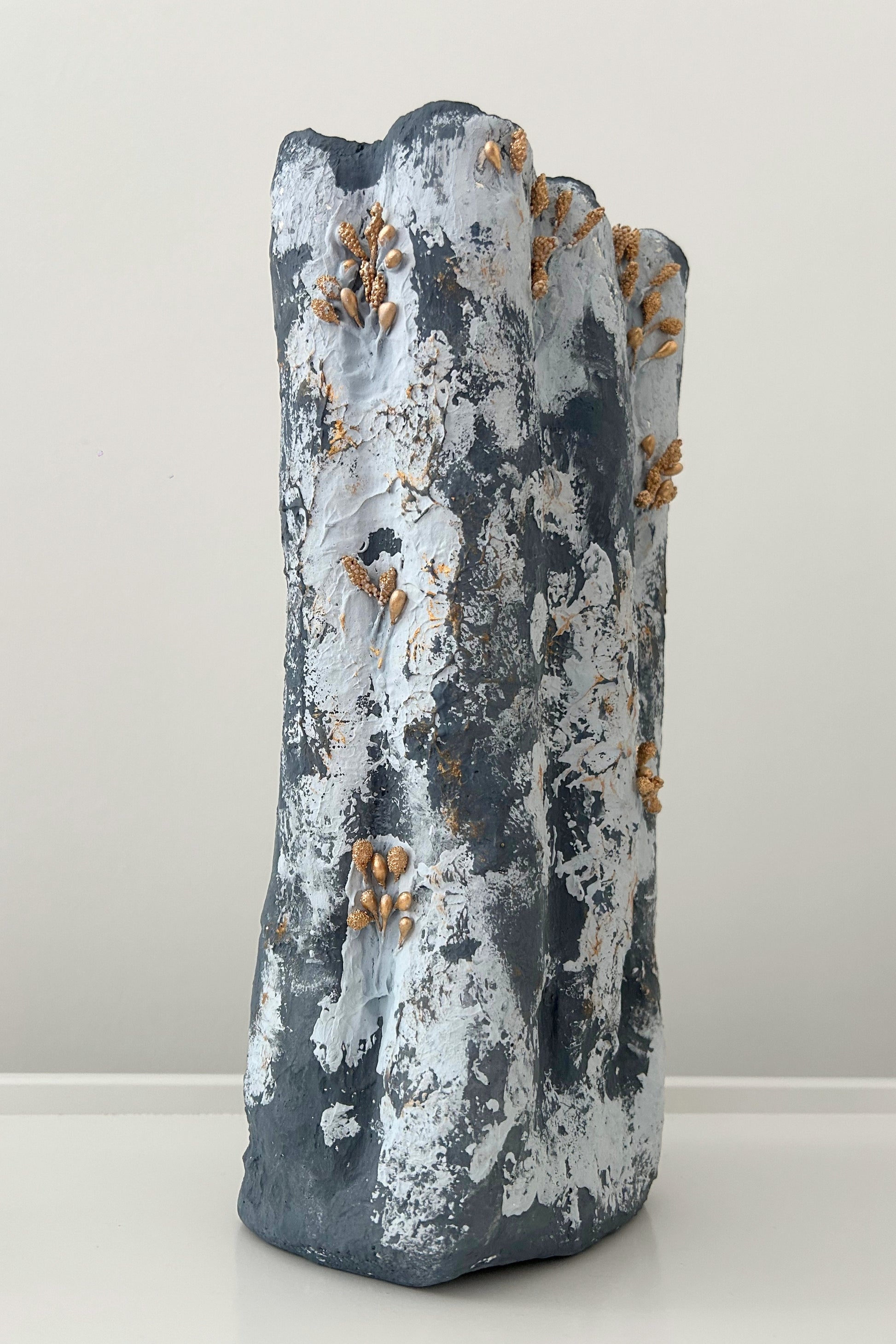Organic tall vase with grey-blue plaster texture and gold accent details, standing against white wall.