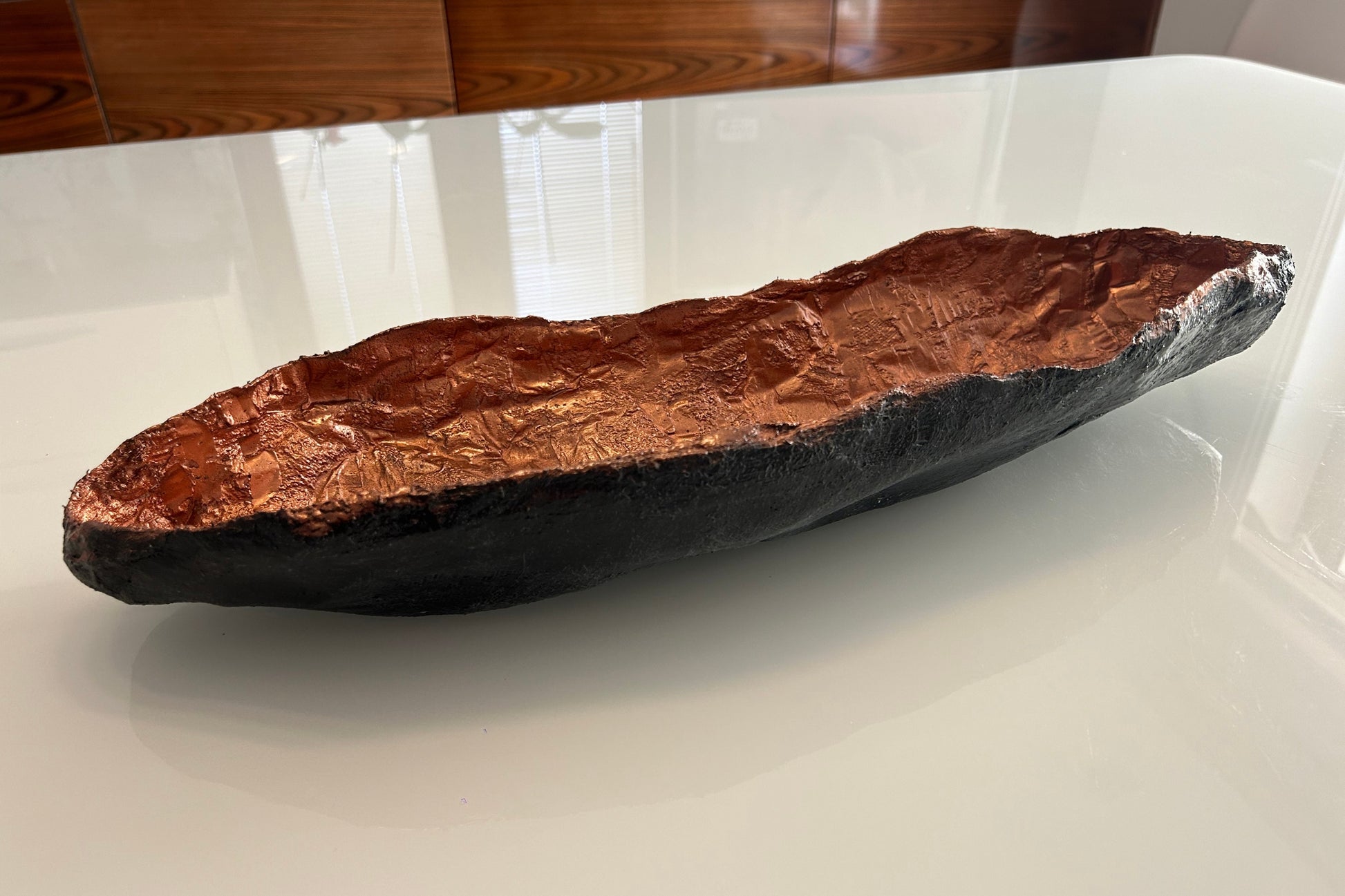 Sculptural long bowl with copper interior and matte black exterior placed on rustic wood.