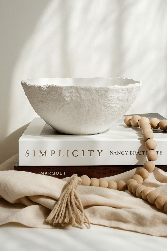 Handmade white textured wabi sabi bowl placed on art books with natural wood beads, styled in soft sunlight.